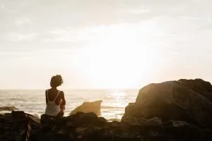 Femme assise de dos sur des rochers, contemplant l’océan et le soleil – symbole d’éveil spirituel et de reconnexion à Soi